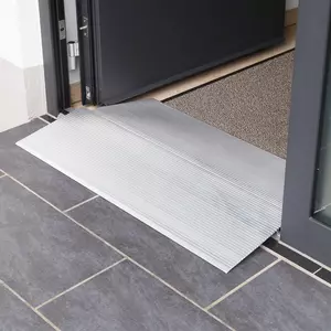 Eine Schwellenbrücke aus Aluminium  liegt an der Schwelle einer Türöffnung über dunklen Bodenfliesen neben einer Fußmatte.