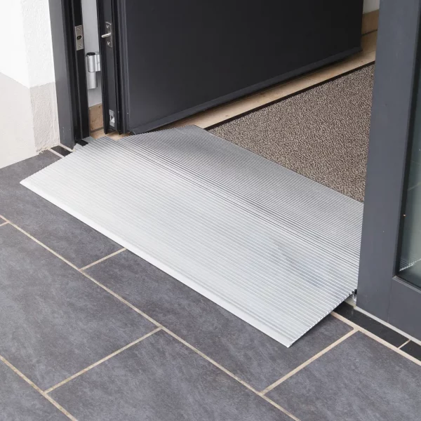 Eine Schwellenbrücke aus Aluminium  liegt an der Schwelle einer Türöffnung über dunklen Bodenfliesen neben einer Fußmatte.