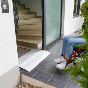 Eine Person im Rollstuhl steht vor einer Schwellenbrücke an der Türschwelle eines Gebäudes