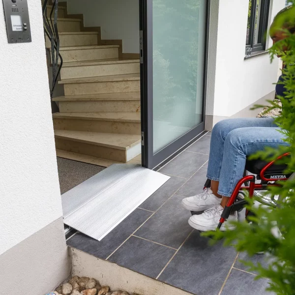 Eine Person im Rollstuhl steht vor einer Schwellenbrücke an der Türschwelle eines Gebäudes
