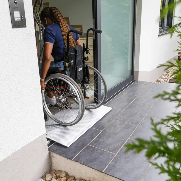 Eine Person im Rollstuhl befährt ein Gebäude über eine Schwellenbrücke neben einer Glastür.