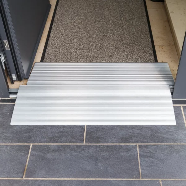Zwei Schwellenbrücken aus Alumimium  für Rollstühle an einem Türeingang mit dunklem Fliesenboden und einer strukturierten Matte im Inneren.