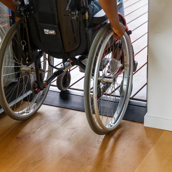 Eine Person im Rollstuhl fährt über einen Schwellenkeil mit niedriger Kante von einem Holzboden im Innenbereich auf eine Terrasse im Außenbereich.