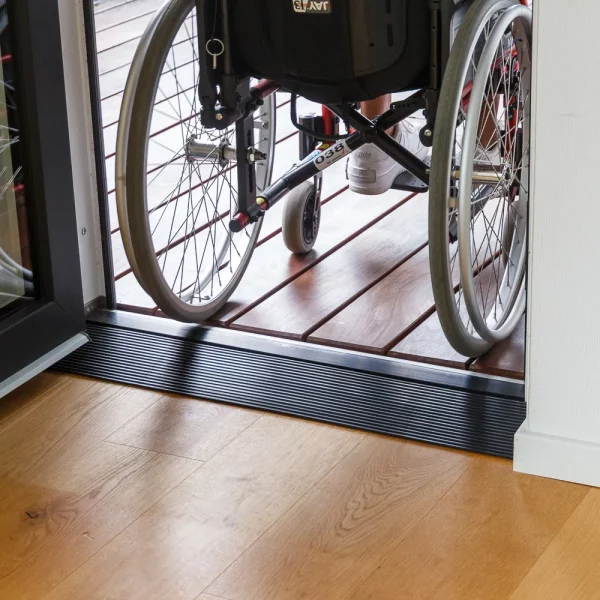 Ein Rollstuhl überquert durch einen Schwellenkeil den Höhenunterschied zwischen einem Holzboden im Innenbereich und einer Terrasse im Freien.