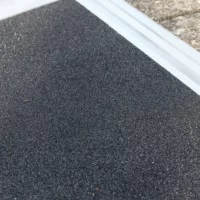 Nahaufnahme der strukturierten schwarzen Fahrfläche einer Rollstuhlrampe neben einer Stein- oder Betonfläche.