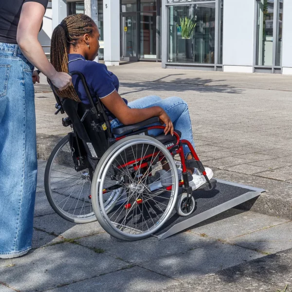 Eine Person im Rollstuhl wird an einem sonnigen Tag im Freien von einer anderen Person eine Bordsteinrampe hinaufgefahren.