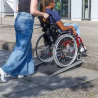 Eine Person schiebt eine andere Person im Rollstuhl über eine tragbare Rampe auf einen Bordstein vor einem Gebäude.