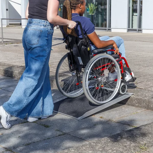 Eine Person schiebt eine andere Person im Rollstuhl über eine tragbare Rampe auf einen Bordstein vor einem Gebäude.
