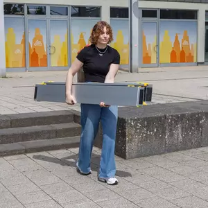 Eine Person mit lockigem Haar trägt eine zusammengeschobene zweiteilige tragbare Rampe im Freien in der Nähe von Betonstufen und einem Gebäude mit orangefarbenen Fensteraufklebern.