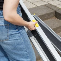 Eine Person in blauen Jeans baut eine tragbare Rampe mit einem gelben Griff in der Nähe von Außentreppen auf.