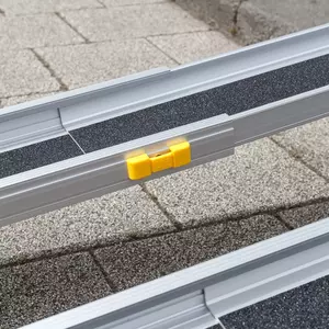 Nahaufnahme der gelben Verriegelung einer aluminiumfarbenen Rollstuhlrampe im Freien