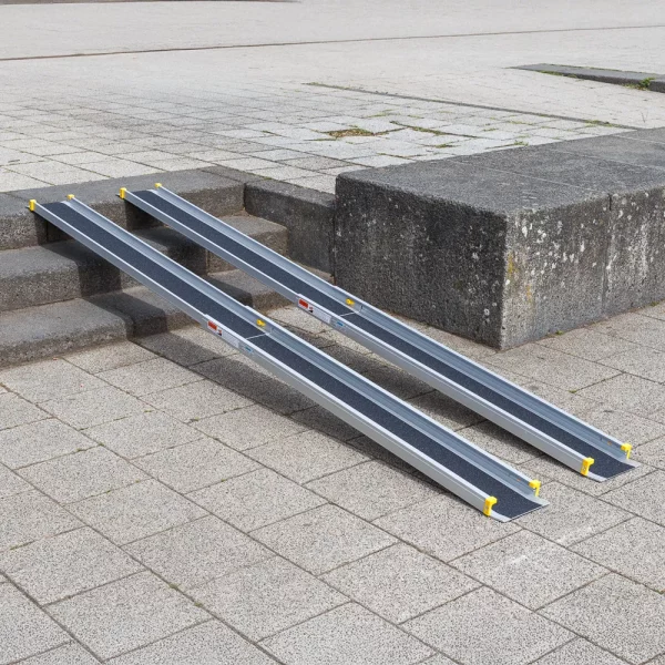 Eine zweiteilige Rollstuhlrampe ist über Steinstufen gelegt und ermöglichen den Zugang von einem niedrigeren zu einem höheren Gehwegniveau.