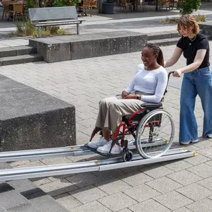 Eine Frau schiebt eine andere Frau im Rollstuhl über eine tragbare Rollstuhlrampe auf einem gepflasterten Außengelände.