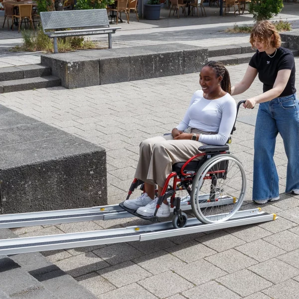 Eine Frau schiebt eine andere Frau im Rollstuhl über eine tragbare Rollstuhlrampe auf einem gepflasterten Außengelände.
