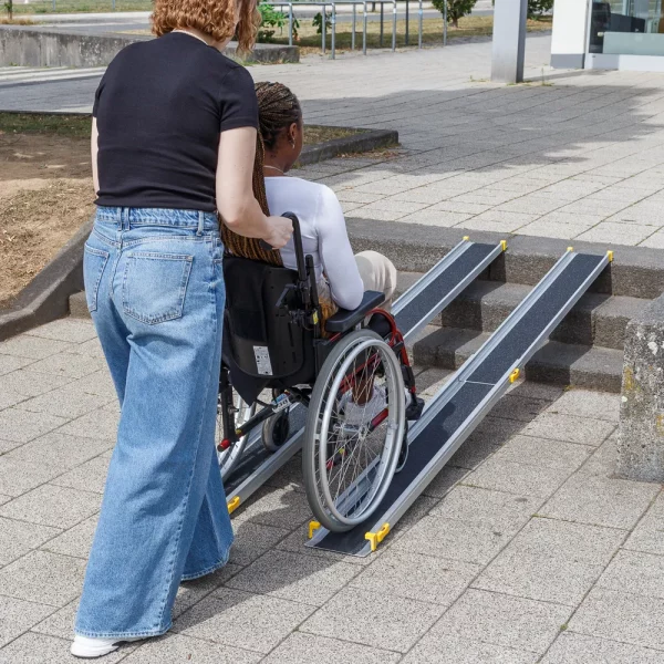 Eine Person schiebt eine andere Person in einem Rollstuhl über tragbare Rollstuhlrampen auf eine Außentreppe.