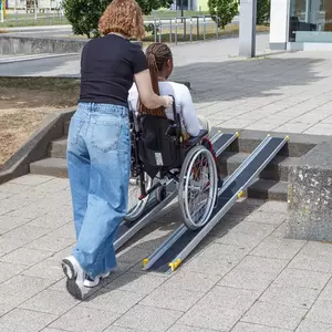 Eine Frau schiebt eine andere Frau im Rollstuhl auf eine tragbare Rampe über eine Außentreppe in der Nähe eines Gebäudes.