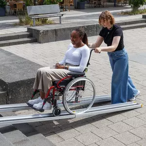 Eine Frau schiebt eine andere Frau im Rollstuhl eine tragbare Rampe auf einer städtischen Treppe hinauf.