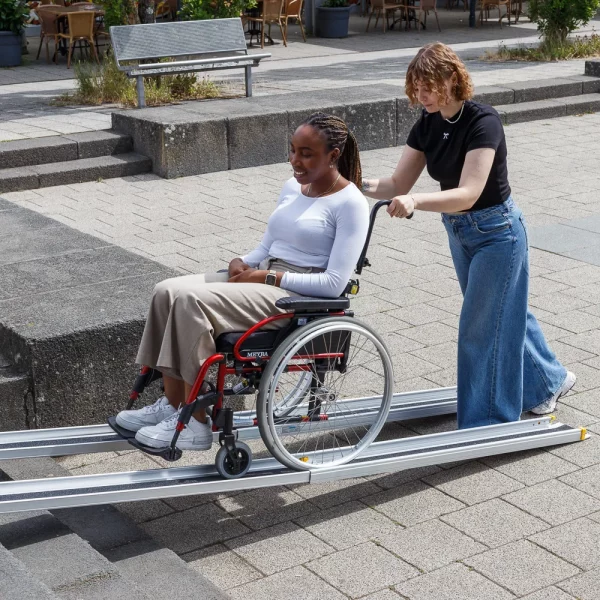 Eine Frau schiebt eine andere Frau im Rollstuhl eine tragbare Rampe auf einer städtischen Treppe hinauf.