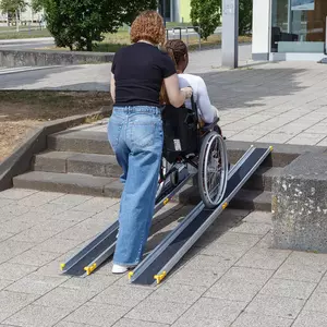 Eine Frau schiebt eine andere Frau im Rollstuhl auf einer tragbaren Rampe über Stufen auf einem gepflasterten Weg.