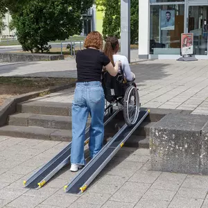Eine Person schiebt eine Rollstuhlfahrerin auf einer tragbaren Rampe über eine Außentreppe in Richtung eines Gebäudeeingangs.