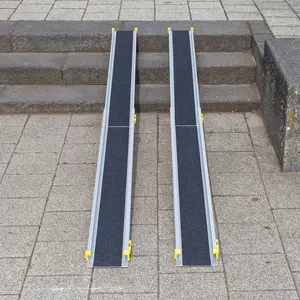 Zwei tragbare Rollstuhlrampen sind über Steinstufen auf einem gefliesten Außenweg angebracht.