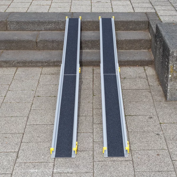 Zwei tragbare Rollstuhlrampen sind über Steinstufen auf einem gefliesten Außenweg angebracht.