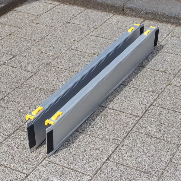 Zwei Rollstuhlrampen aus Aluminium mit gelben Griffen sind zusammengeschoben auf eine gepflasterte Außenfläche gestellt.