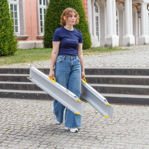 Eine Person in Jeans und marineblauem Shirt trägt zwei Rollstuhlrampen-Schienen im Freien in die Nähe von Steinstufen und einem Gebäude mit Säulen.