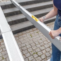 Eine Person stellt eine Rollstuhlrampe aus Aluminium auf die Steinstufen im Freien und hält die Rampe mit beiden Händen fest.