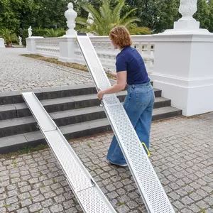 Eine Person in Jeans stellt eine tragbare Aluminiumrampe über Steinstufen im Freien in der Nähe eines weißen Geländers auf.