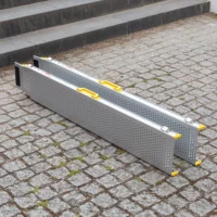 Zwei tragbare Rollstuhlrampen-Schienen mit gelben Griffen sind nebeneinander auf einem Kopfsteinpflaster in der Nähe einiger Stufen aufgestellt.