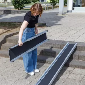 Eine Person in Jeans und schwarzem Shirt stellt eine tragbare Rampe über einer Außentreppe auf einem gepflasterten Gehweg auf.