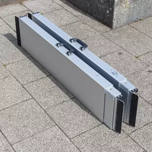 Eine zweiteilige zusammengelegte Aluminiumrampe mit schwarzen Griffen steht auf einer gepflasterten Außenfläche.