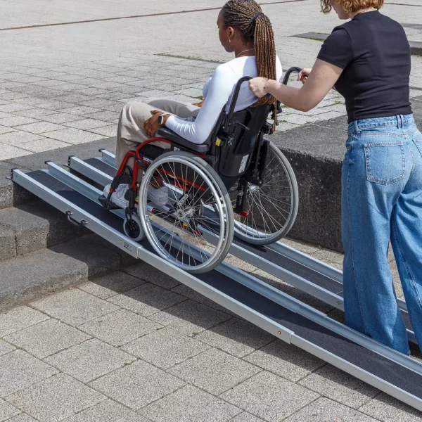 Eine Person schiebt eine andere Person im Rollstuhl eine tragbare Rampe über Betonstufen im Freien hinauf.